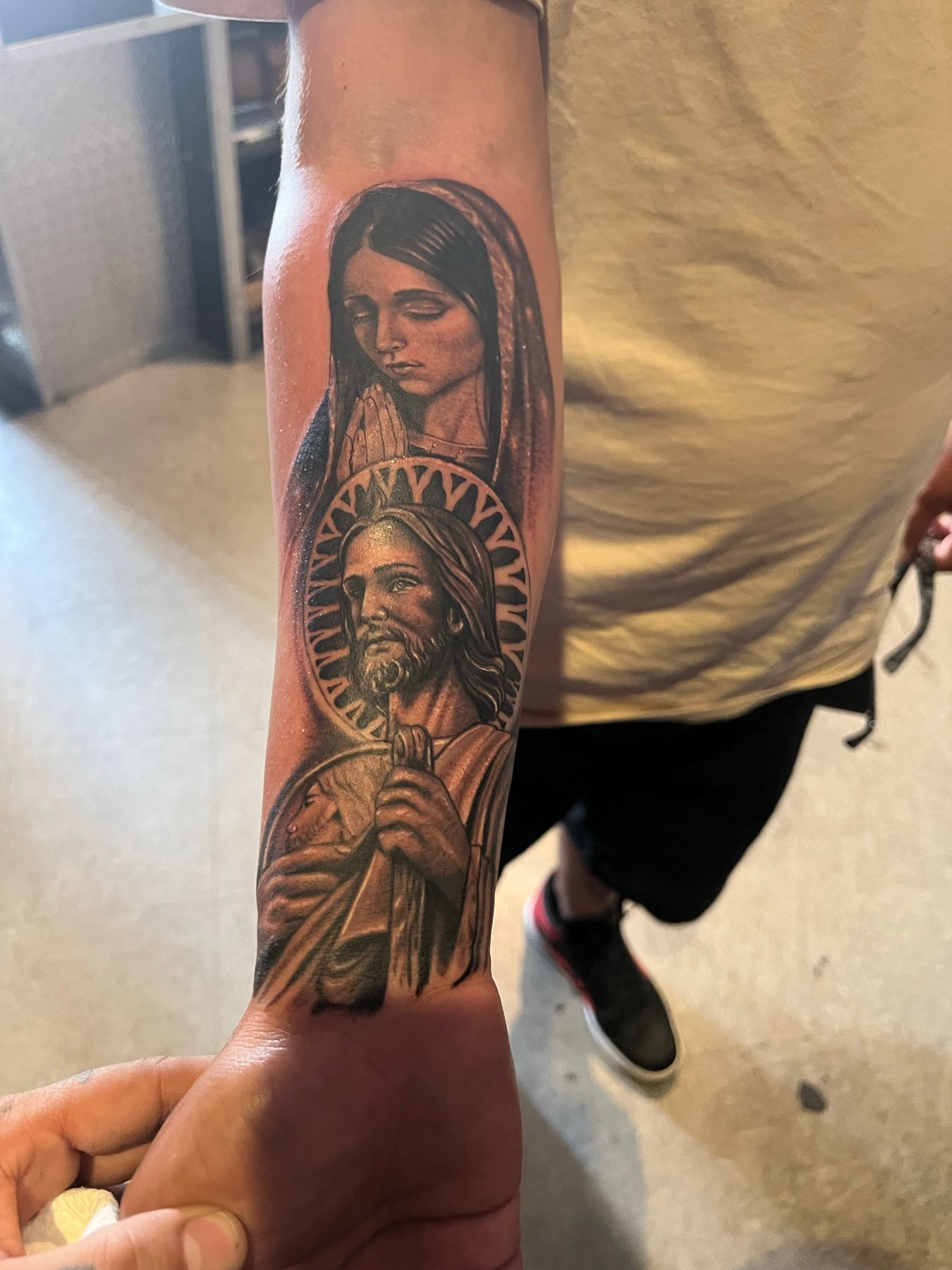 Reworked black and grey religious forearm tattoo of two saints with a radiant halo, by American Ink in Sherman Oaks.