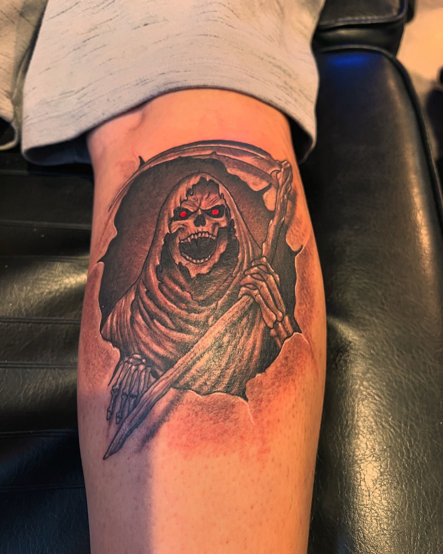 Black and grey tattoo of a hooded grim reaper with red eyes holding a scythe on a calf, by American Ink in Sherman Oaks.