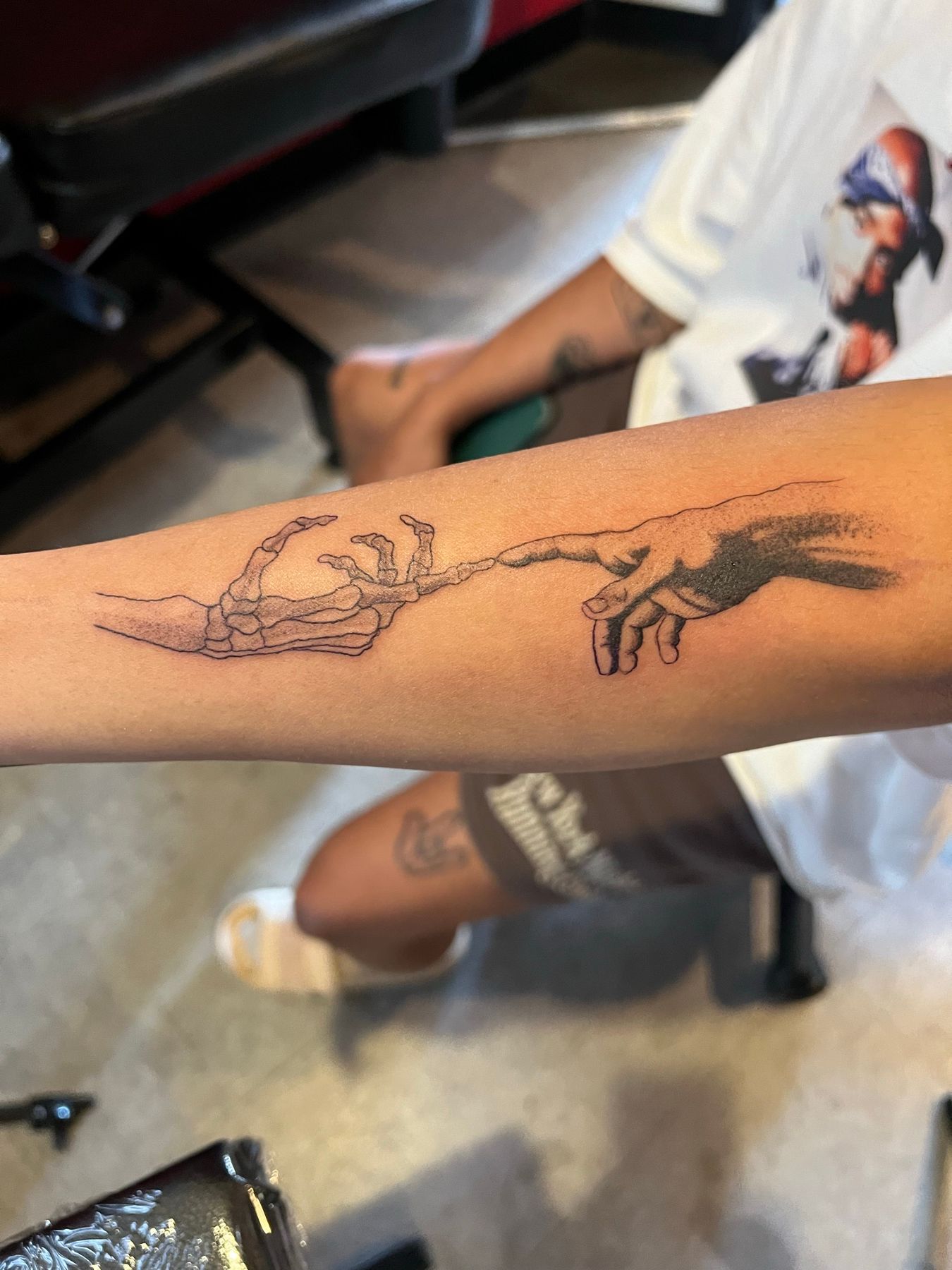 Fine line and dotwork tattoo of a human hand reaching toward a skeletal hand in a Creation of Adam composition on a forearm, by American Ink in Sherman Oaks.