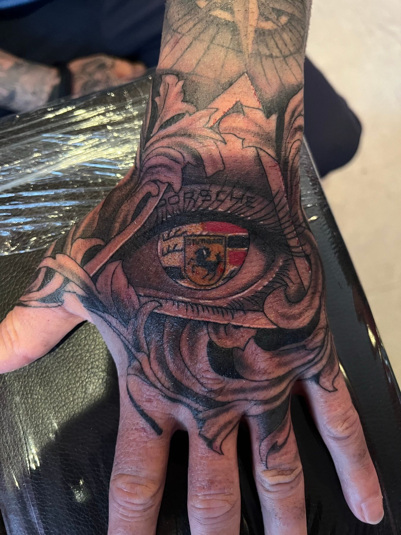 Black and grey tattoo of a color Porsche crest framed by baroque filigree on the back of a hand, by American Ink in Sherman Oaks.