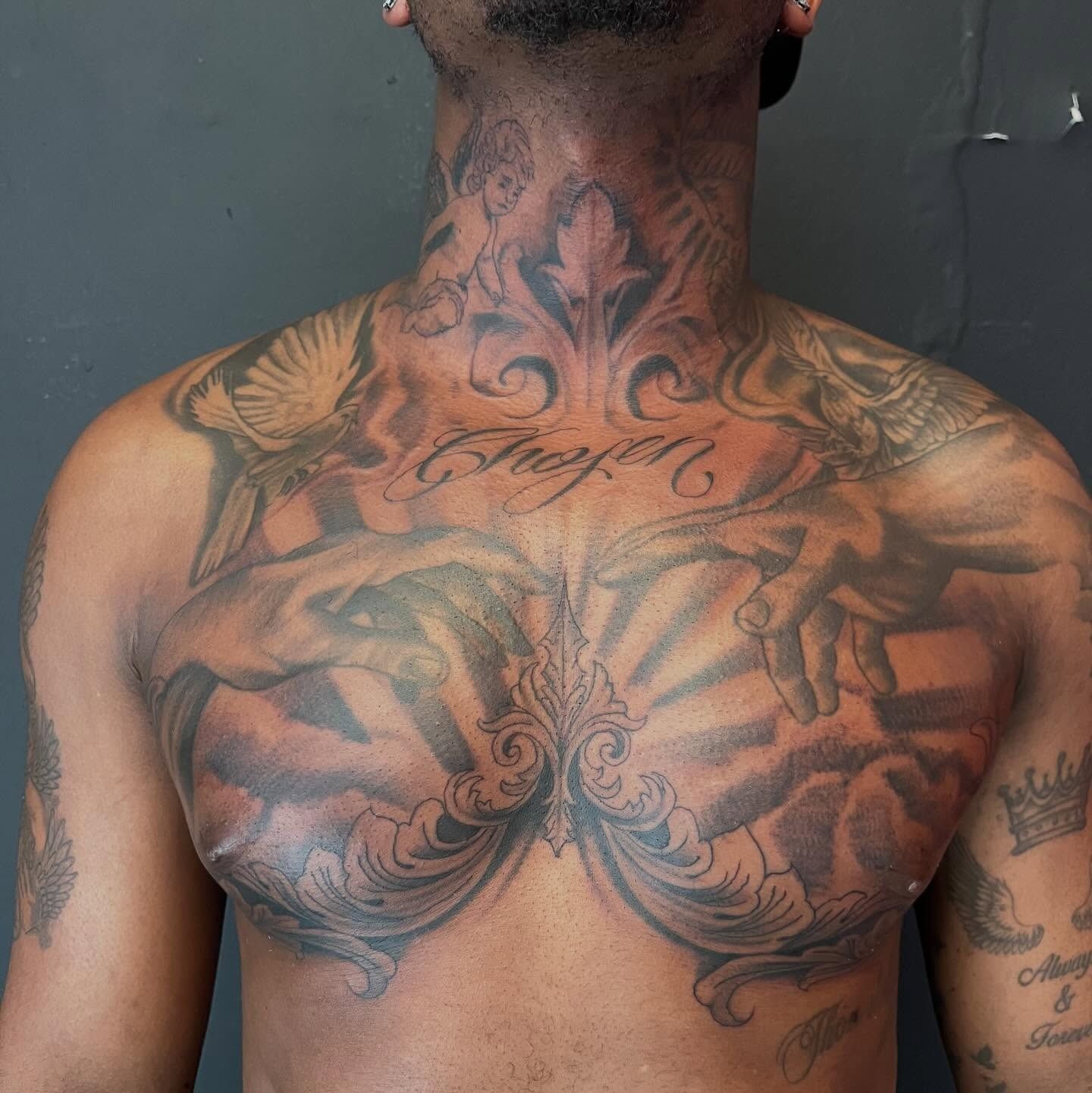 Full chest and neck black and grey tattoo with cherub, doves, reaching hands, sun rays, and Chosen script lettering, by Josue at American Ink in Sherman Oaks.