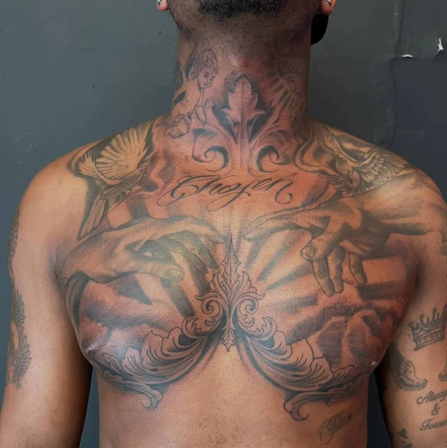 Full chest and neck black and grey tattoo with cherub, doves, reaching hands, sun rays, and Chosen script lettering, by Josue at American Ink in Sherman Oaks.