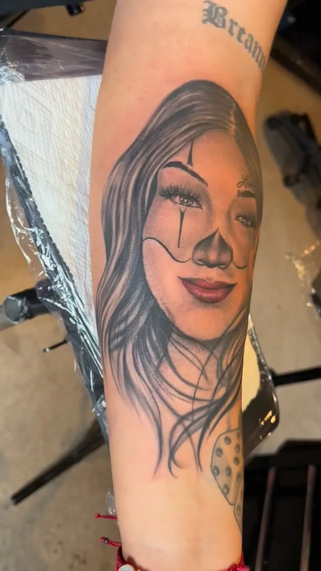 Black and grey Chicano payasa forearm portrait with clown face paint, flowing hair, and red lips, tattooed by Josue at American Ink in Sherman Oaks.