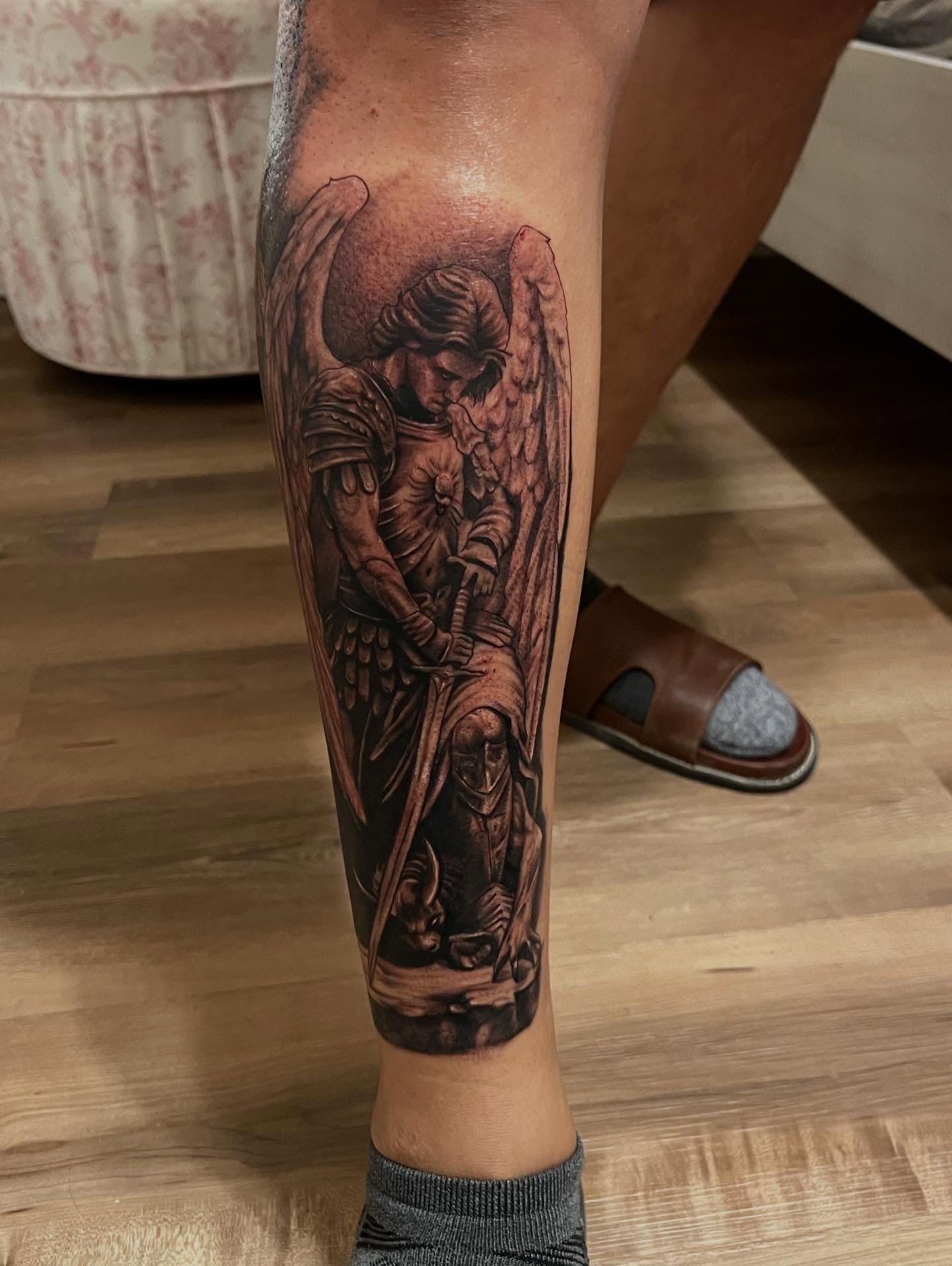 Black and grey calf tattoo of Archangel Michael in winged armor lowering his sword over a defeated demon, by Josue at American Ink in Sherman Oaks.