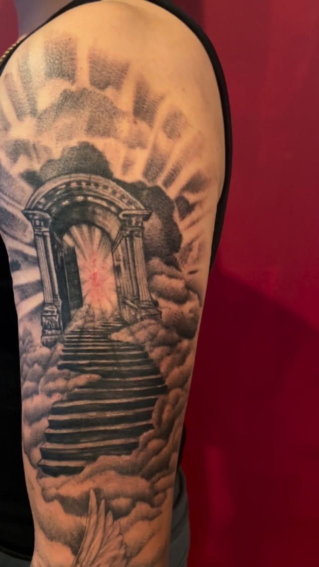 Black and grey half-sleeve of a stairway rising through clouds to an arched gate with light rays, tattooed by Josue at American Ink in Sherman Oaks.
