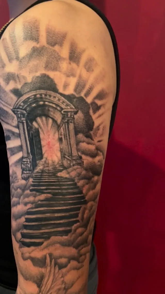 Black and grey half-sleeve of a stairway rising through clouds to an arched gate with light rays, tattooed by Josue at American Ink in Sherman Oaks.
