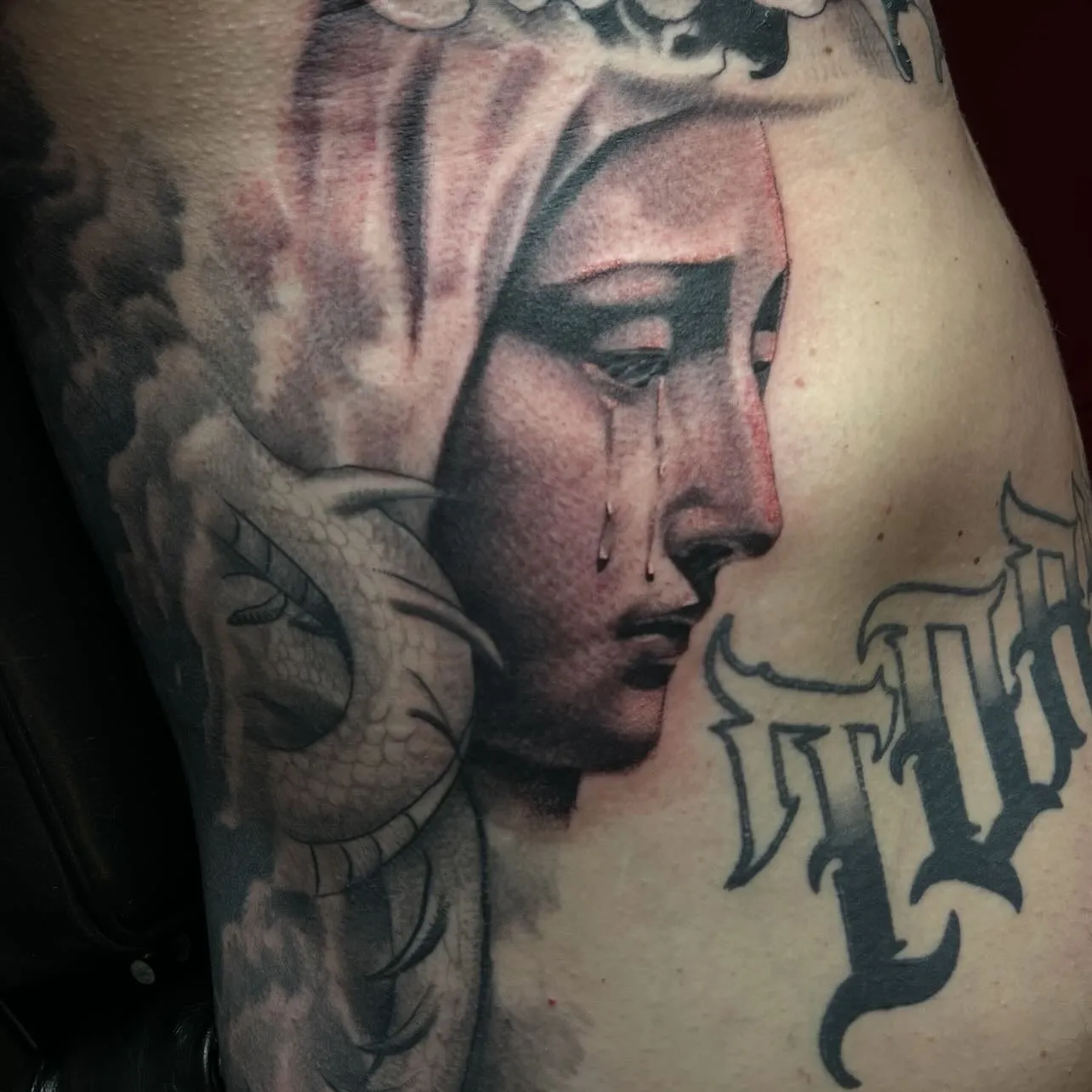 Close-up of a black and grey rib tattoo of a weeping Madonna in profile with a dragon and gothic lettering, by Josue at American Ink in Sherman Oaks.