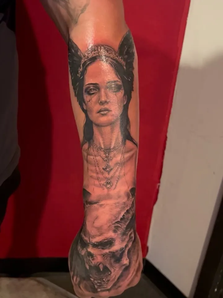 Dark black and grey forearm tattoo of a winged woman in a jeweled crown with layered necklaces above a skull, by Josue at American Ink in Sherman Oaks.