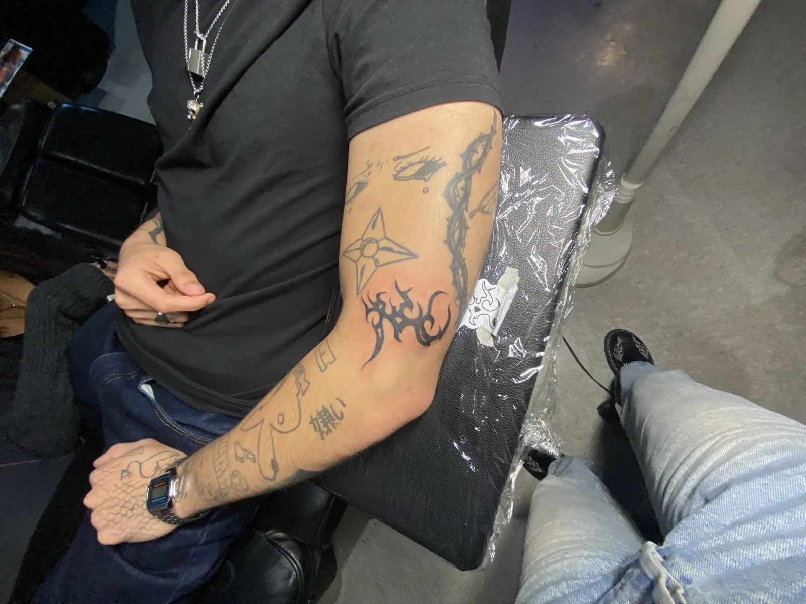 Fine-line patchwork forearm tattoos by Knives — a throwing star, gothic lettering, an eyes motif, and a small fish across the hand, in black ink at American Ink in Sherman Oaks.