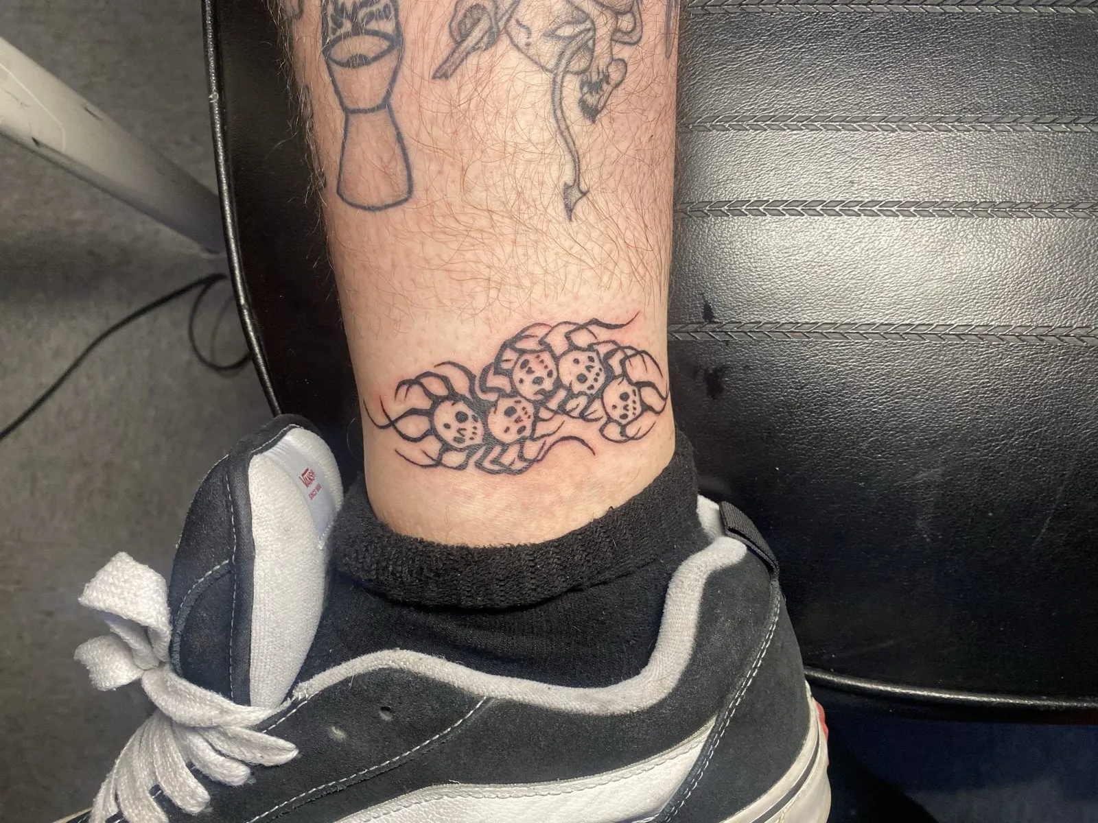 Fine-line ankle-band tattoo by Knives of stylized skulls woven with tribal flame flourishes, with smaller fine-line pieces up the shin — tattoo work at American Ink in Sherman Oaks.
