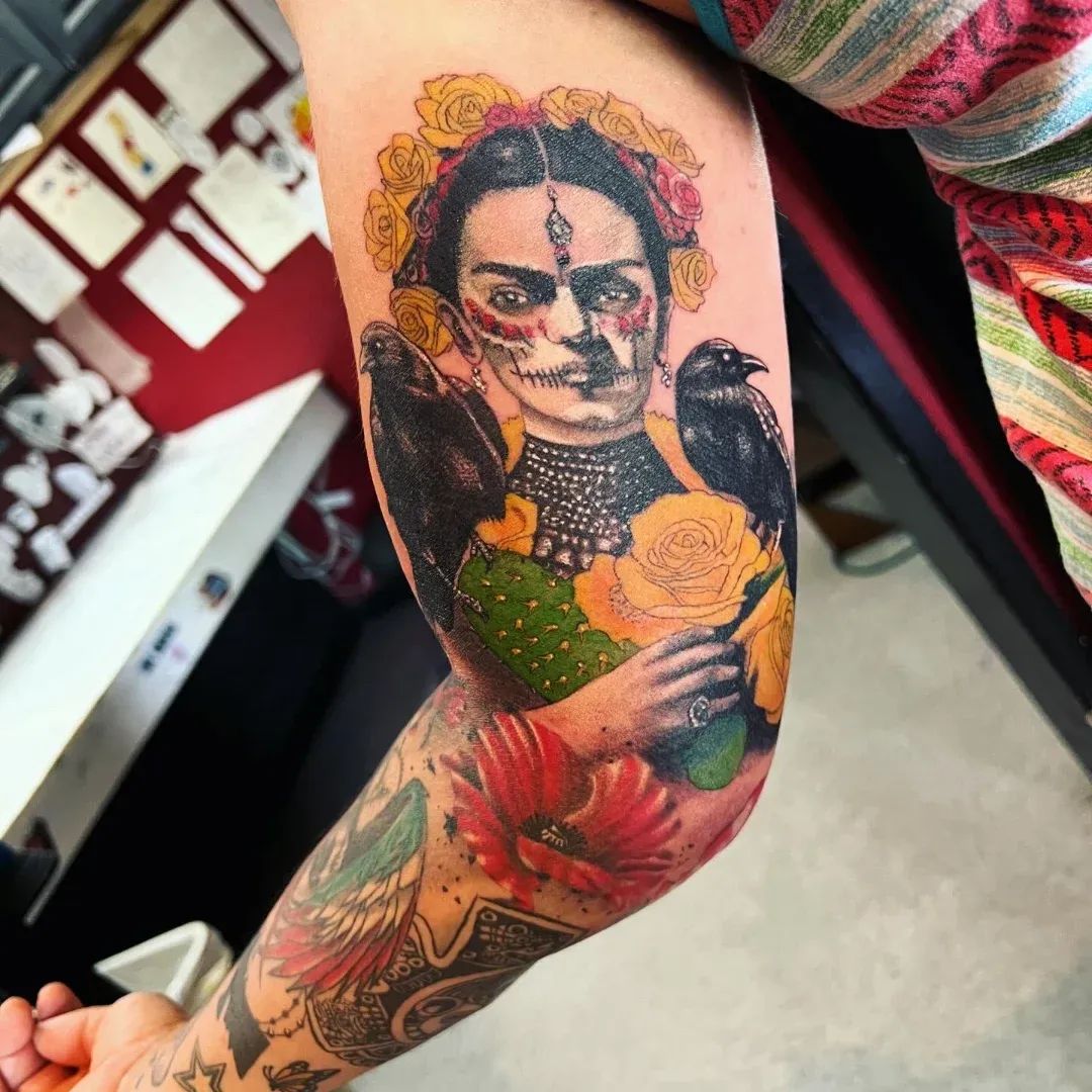 Color portrait tattoo of Frida Kahlo with sugar-skull makeup, yellow roses, ravens, and a cactus on an upper arm, from American Ink Tattoos in Sherman Oaks.