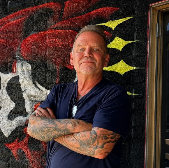 Miguel, owner and piercer at American Ink Tattoo in Sherman Oaks, standing with arms crossed in front of the shop mural.