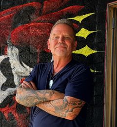 Miguel, owner and piercer at American Ink Tattoo in Sherman Oaks, standing with arms crossed in front of the shop mural.
