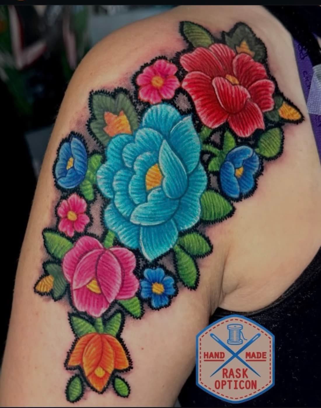 Color embroidery patch-style shoulder tattoo of a stitched floral bouquet in blue, red, pink, and orange, tattooed by Rask Opticon at American Ink in Sherman Oaks.