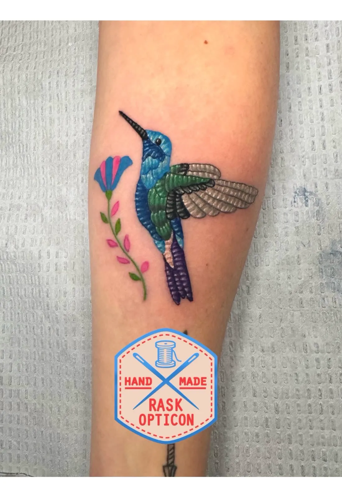 Embroidery patch-style hummingbird and flower tattoo on the forearm with Rask Opticon's spool-and-needle maker's mark, at American Ink in Sherman Oaks.