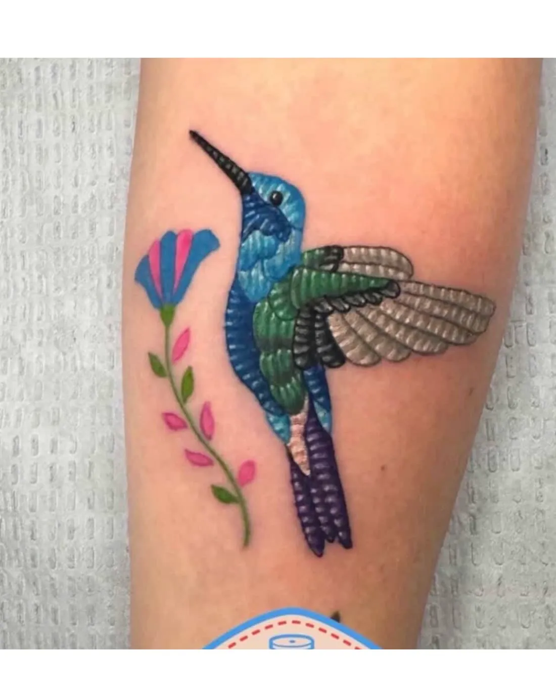 Embroidery patch-style tattoo of a blue and green hummingbird beside a pink flower on the forearm, by Rask Opticon at American Ink in Sherman Oaks.