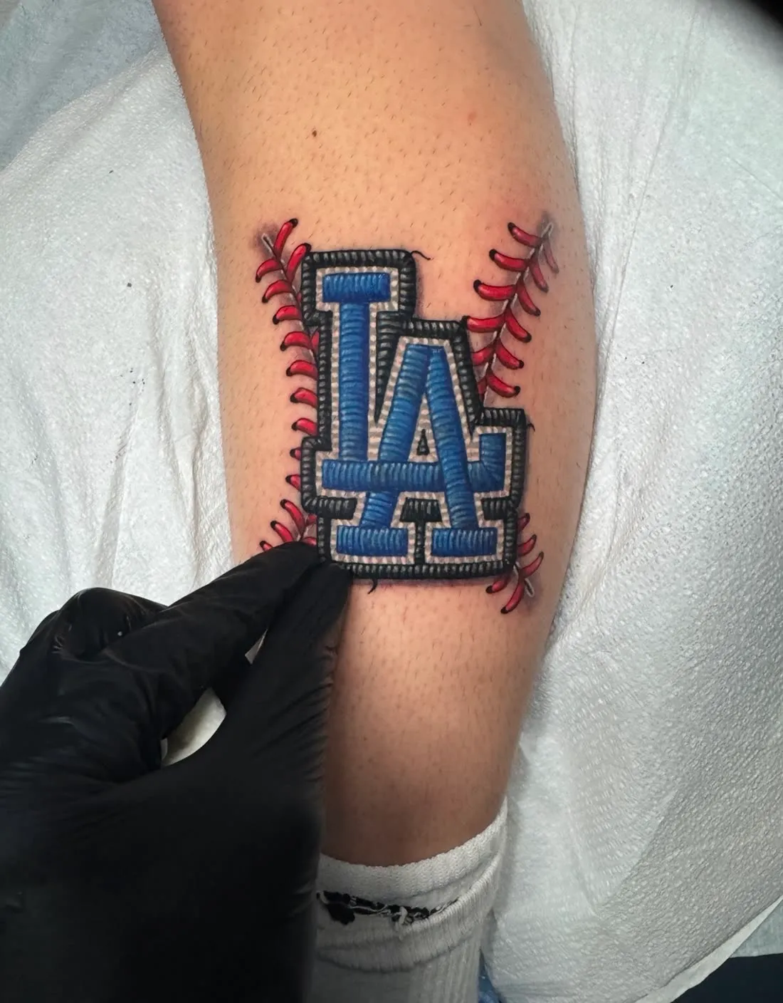 Embroidery patch-style tattoo of the LA Dodgers logo framed by red baseball stitching, by Rask Opticon at American Ink in Sherman Oaks.