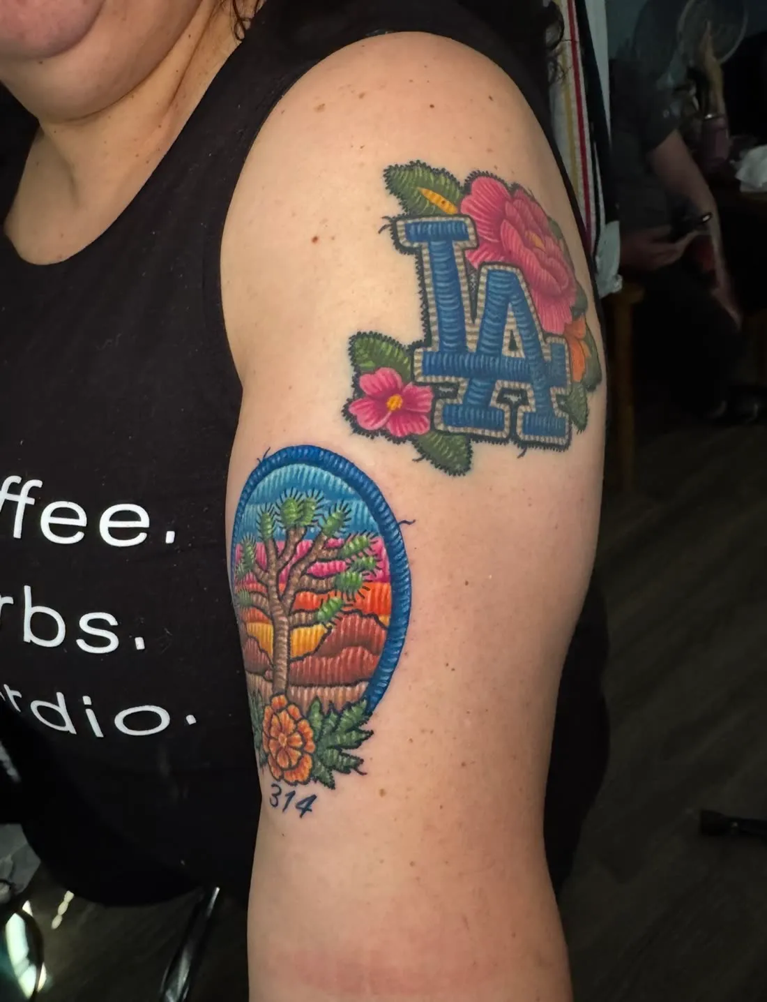 Two embroidery patch-style tattoos on the upper arm — an LA Dodgers logo with flowers and a Joshua Tree desert scene — by Rask Opticon at American Ink in Sherman Oaks.