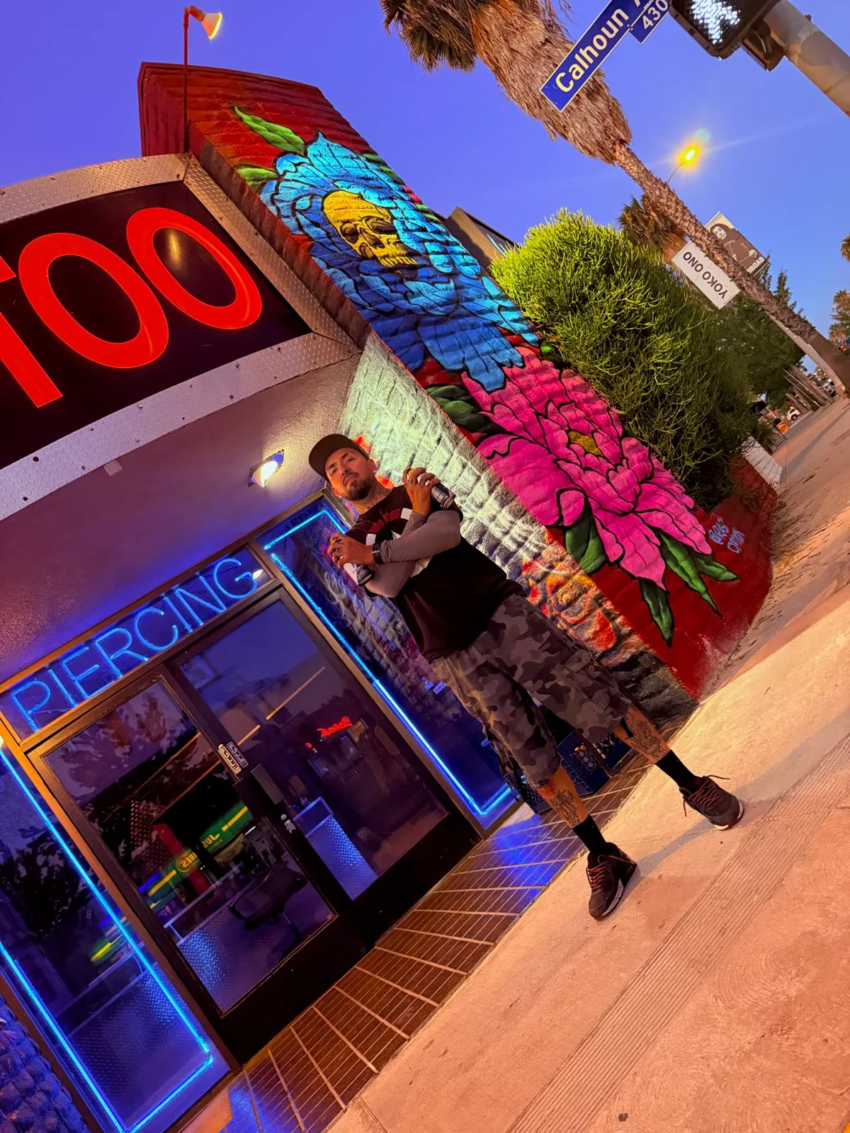 Rask Opticon holding spray cans in front of the American Ink storefront mural and neon piercing sign in Sherman Oaks at dusk.