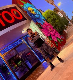 Rask Opticon holding spray cans in front of the American Ink storefront mural and neon piercing sign in Sherman Oaks at dusk.