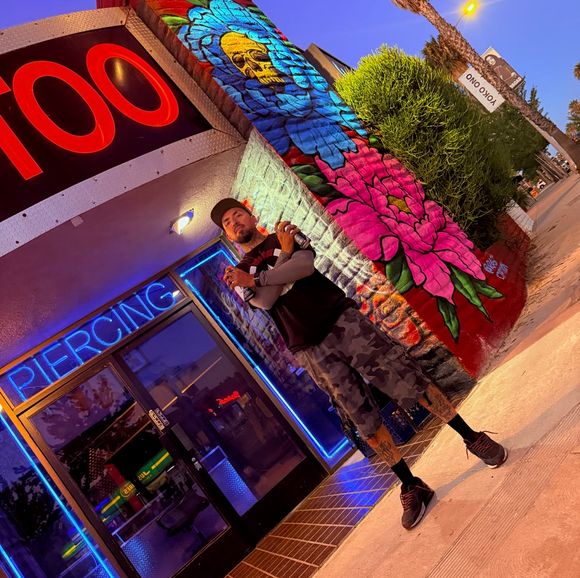 Rask Opticon holding spray cans in front of the American Ink storefront mural and neon piercing sign in Sherman Oaks at dusk.
